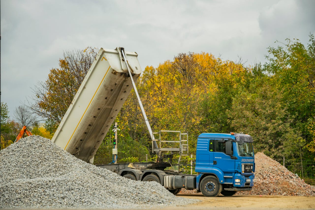 Blue dump truck unloading gravel at an outdoor construction site surrounded by autumn trees.