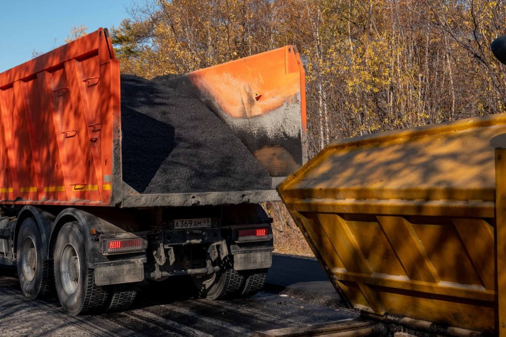 A dump truck unloading asphalt into a construction bin on a sunny autumn day.